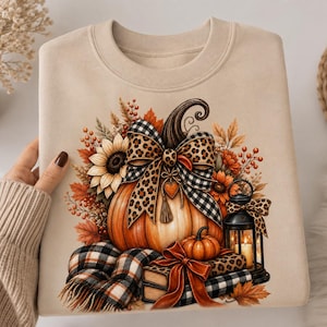 Op de afbeelding: Een beige sweater met ronde hals voorzien van een grafische print van een oranje pompoen versierd met een strik in luipaardprint en zwart-wit ruitpatroon, omringd door zonnebloemen, herfstbladeren, een lantaarn, boeken en een sjaal.