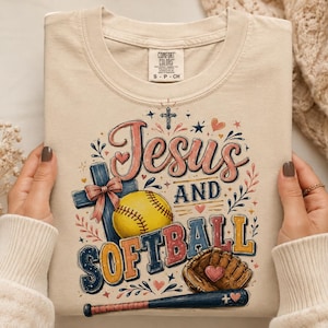 Op de afbeelding: Een beige t-shirt met een grafische print met de tekst Jesus and Softball. Het ontwerp bevat een blauw kruis met een roze strik, een gele softbal, een bruine honkbalhandschoen met een roze hart en een blauwe honkbalknuppel.