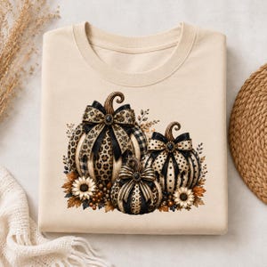 Op de afbeelding: Een beige T-shirt met ronde hals en een grafische print van drie pompoenen. De pompoenen zijn versierd met zwarte en beige luipaardprints, ruitpatronen en stippen, elk bekroond met een grote zwarte strik en omringd door zonnebloemen.