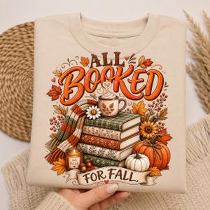 Op de afbeelding: Een beige sweater met ronde hals en een grafische print met de tekst ALL BOOKED FOR FALL. Het ontwerp bevat een stapel van vier boeken, een dampende mok, een gestreepte sjaal, een kaars, pompoenen en herfstbladeren in oranje, rood en groen.
