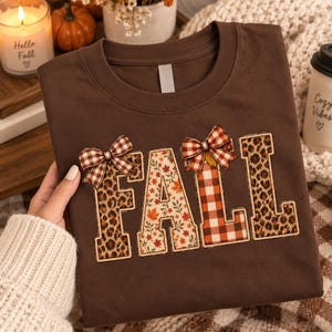 Op de afbeelding: Een donkerbruin t-shirt met het woord FALL in grote blokletters. De letters zijn gevuld met luipaardprint, herfstbladpatronen en oranje met witte ruiten. Twee kleine geruite strikjes zijn aan de bovenkant van de letters bevestigd.