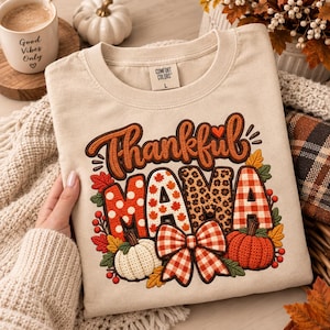 Op de afbeelding: Een beige T-shirt met ronde hals en de tekst Thankful MAMA in bruin, waarbij de letters zijn gevuld met stippen, esdoornbladeren, luipaardprint en ruitpatronen. Het ontwerp bevat een strik met ruitjes, twee pompoenen en herfstbladeren en bessen.