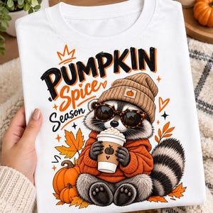 Op de afbeelding: Een wit t-shirt met een afbeelding van een wasbeer die een bruine muts en een schildpad-zonnebril draagt en een koffiebeker vasthoudt. De tekst luidt PUMPKIN Spice Season. Het ontwerp bevat oranje pompoenen en herfstbladeren.