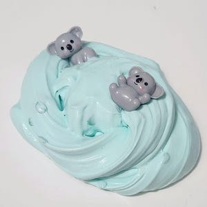 May include: A mound of light blue, swirled, textured slime featuring two small, grey, glossy koala bear figurines embedded into the surface. The koalas have black eyes, black noses, and small pink cheeks, resting on the soft, pliable material.