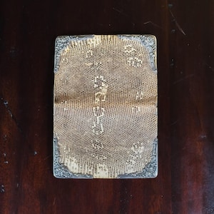 May include: A rectangular card case featuring a textured, tan-colored snakeskin-like pattern on the exterior. The four corners are reinforced with ornate, silver-toned metal accents that display intricate, embossed floral and scrollwork detailing.