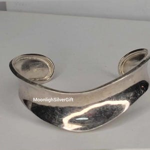 May include: A wide, polished silver cuff bracelet with a smooth, undulating wave-like shape and a curved open design. The surface reflects light, highlighting its metallic sheen and fluid form. The text MoonlighSilverGift is visible on the surface.