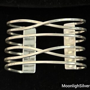 May include: A silver-toned cuff bracelet featuring five horizontal bands that intersect in a crisscross pattern at the center. The bands are connected by two vertical rectangular bars on either side, creating an open, cage-like structure.