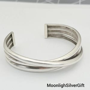 Könnte beinhalten: Ein silberfarbenes Armband in Manschettenform mit einem mehrsträngigen Design aus drei überlappenden Bändern, die an den Enden zusammenlaufen. Das Metall hat eine glatte, polierte Oberfläche und eine offene Rückseite. Text MoonlighSilverGift ist sichtbar.