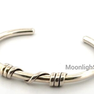 Puede incluir: Una pulsera tipo brazalete de plata con una banda lisa y redondeada que presenta dos secciones decorativas de alambre envuelto tres veces. El metal tiene un acabado pulido y el texto MoonlighSilverGift es visible en el lado derecho.