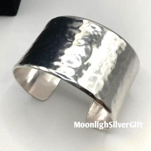 Könnte beinhalten: Ein breiter Silberarmreif mit einer gehämmerten, texturierten Oberfläche, die das Licht reflektiert. Das Metall hat ein poliertes, glänzendes Finish und ein offenes Design. Der Text MoonlighSilverGift ist unten rechts im Bild zu sehen.