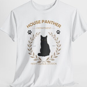 Può includere: Una t-shirt bianca a maniche corte con la grafica di un gatto nero centrato all'interno di una corona d'alloro color marrone chiaro. Il testo recita HOUSE PANTHER DEPARTMENT, EST 1987 e NIGHT PATROL DIVISION, con due piccole impronte.