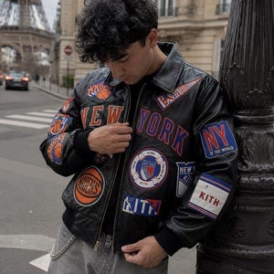 May include: A black leather varsity jacket featuring various embroidered patches including New York Knicks logos, Kith branding, and New York Rangers emblems. The jacket has a front zipper closure, a classic collar, and black ribbed cuffs and hem.