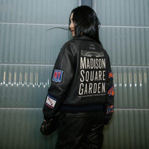 May include: A black leather varsity jacket featuring the text Madison Square Garden, The World's Most Famous Arena, and various patches including NY To The World, Kith, and Knicks logos on the back and sleeves with ribbed cuffs and hem.