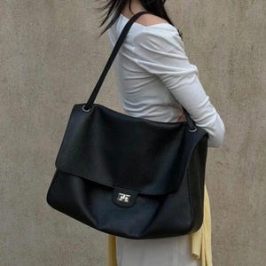 Puede incluir: Un bolso de hombro grande de color negro con superficie texturizada, que presenta una solapa ancha plegable y un cierre giratorio rectangular de tono plateado. El bolso tiene una sola correa larga para el hombro sujeta a los lados con anillas metálicas plateadas.