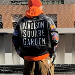 May include: A black leather jacket featuring the text MADISON SQUARE GARDEN THE WORLD'S MOST FAMOUS ARENA on the back, paired with an orange hooded sweatshirt, an orange beanie, and brown cargo pants with large side pockets and visible stitching.