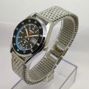 May include: A silver-tone Seiko 5 automatic wristwatch with a black dial featuring 24-hour markings, a day-date window, and a blue accent ring. The watch has a metallic link bracelet with a textured, woven-style pattern and a polished silver case.