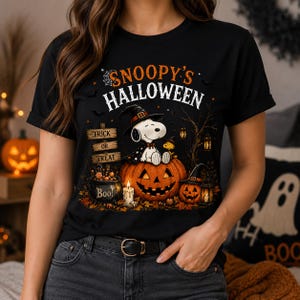 May include: A black t-shirt featuring a graphic of Snoopy wearing a witch hat, sitting on a large jack-o'-lantern. The design includes the text "SNOOPY'S HALLOWEEN", a "TRICK OR TREAT" sign, a cauldron labeled "BOO!", a candle, and a smaller pumpkin.