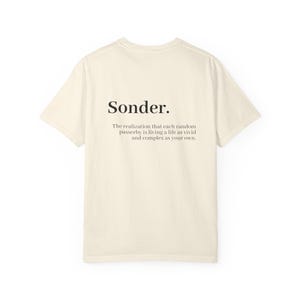 May include: A cream-colored short-sleeve t-shirt featuring the word Sonder in a bold black font, followed by the definition: The realization that each random passerby is living a life as vivid and complex as your own, printed in a smaller black font.