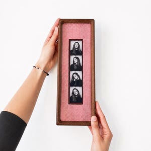 Pode incluir: Uma moldura retangular de madeira escura contendo uma tira vertical de quatro retratos em preto e branco de uma pessoa. O fundo dentro da moldura é um tecido com listras finas vermelhas e brancas. A moldura tem uma borda profunda.