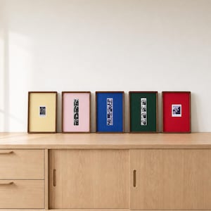 Op de afbeelding: Vijf rechthoekige houten lijsten op een rij, elk met een andersgekleurde passe-partout: geel, roze, blauw, groen en rood. De lijsten bevatten kleine zwart-wit fotostrips of losse vierkante foto's die in het midden van de matten zijn geplaatst.