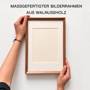 May include: A rectangular walnut wood picture frame with a cream-colored mat board. The text MASSGEFERTIGTER BILDERRAHMEN AUS WALNUSSHOLZ is printed in black above the frame. The frame has a smooth finish and a simple, clean design.