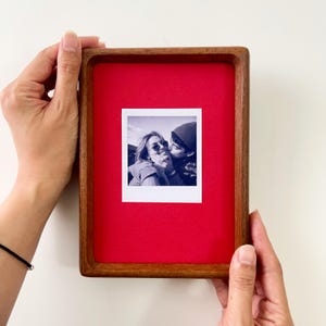 Pode incluir: Uma moldura retangular de madeira com um fundo de feltro vermelho vibrante. Centrada na moldura está uma fotografia instantânea quadrada a preto e branco que mostra duas pessoas, uma com óculos de sol e outra com um gorro, num abraço.