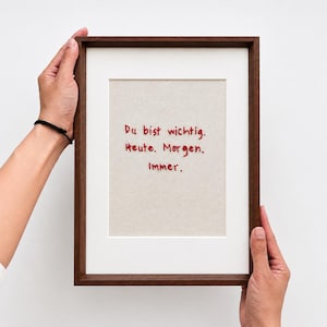 May include: A rectangular dark wood frame containing a light beige fabric insert with red embroidered text that reads: Du bist wichtig. Heute. Morgen. Immer. The frame features a white mat border surrounding the central embroidered fabric panel.