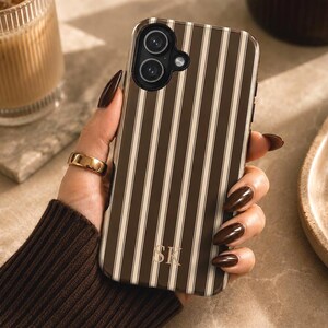 May include: A brown and white vertically striped phone case featuring a dual-lens camera cutout at the top left and the gold-colored monogram letters SK printed near the bottom center. The case has a smooth finish and a rectangular shape.