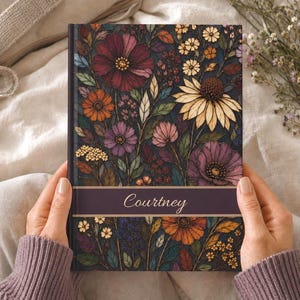 Peut inclure: Un carnet à couverture rigide présentant un fond sombre avec un motif floral vibrant composé de fleurs rouges, orange, violettes et jaunes ainsi que de feuilles vertes. Une bande horizontale violette au centre affiche le prénom Courtney.