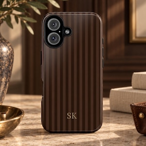 May include: A dark brown smartphone case featuring a vertical pinstripe pattern and the gold-colored initials SK printed near the bottom. The case has a cutout for a dual-lens camera system and a flash, with a smooth, glossy finish throughout.