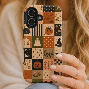 May include: A rectangular phone case featuring a patchwork grid design with various autumn and Halloween-themed illustrations including pumpkins, ghosts, a black cat, a witch hat, spider webs, stars, flowers, and bows in orange, black, and cream.