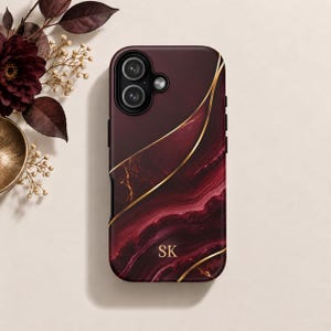 May include: A burgundy phone case featuring a dark red marble pattern with gold-colored veins and curved lines. The case includes a camera cutout and the gold-colored initials SK printed near the bottom center of the back surface.
