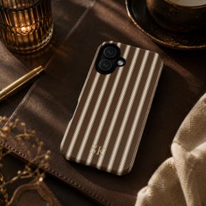 K&ouml;nnte beinhalten: Eine Smartphone-H&uuml;lle mit vertikalem Streifenmuster in Dunkelbraun und Creme. Die H&uuml;lle verf&uuml;gt &uuml;ber einen Kameraausschnitt und die goldfarbenen Initialen SK, die in einer Serifenschrift unten mittig auf der R&uuml;ckseite aufgedruckt sind.