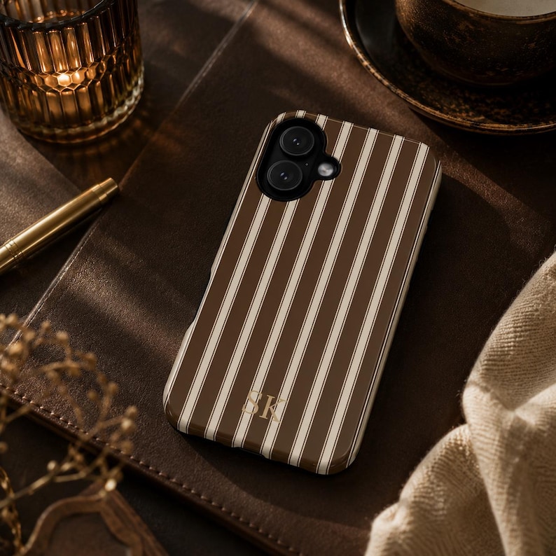 K&ouml;nnte beinhalten: Eine Smartphone-H&uuml;lle mit vertikalem Streifenmuster in Dunkelbraun und Creme. Die H&uuml;lle verf&uuml;gt &uuml;ber einen Kameraausschnitt und die goldfarbenen Initialen SK, die in einer Serifenschrift unten mittig auf der R&uuml;ckseite aufgedruckt sind.
