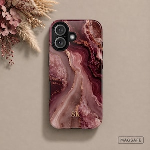 May include: A phone case featuring a deep burgundy and mauve marbled pattern with gold-toned veining and the initials SK printed in gold. The case has a dual-lens camera cutout and a MagSafe label in the corner, showcasing a smooth finish.