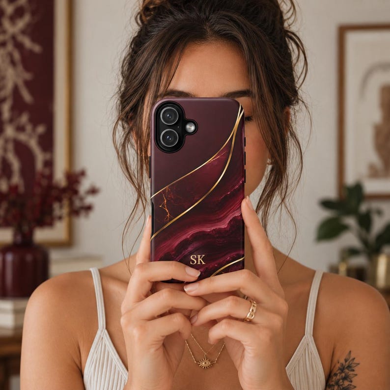 May include: A dark burgundy phone case featuring a swirling marble pattern with thin, elegant gold lines running diagonally across the surface. The bottom center of the case displays the gold-colored initials SK in a clean, serif font.