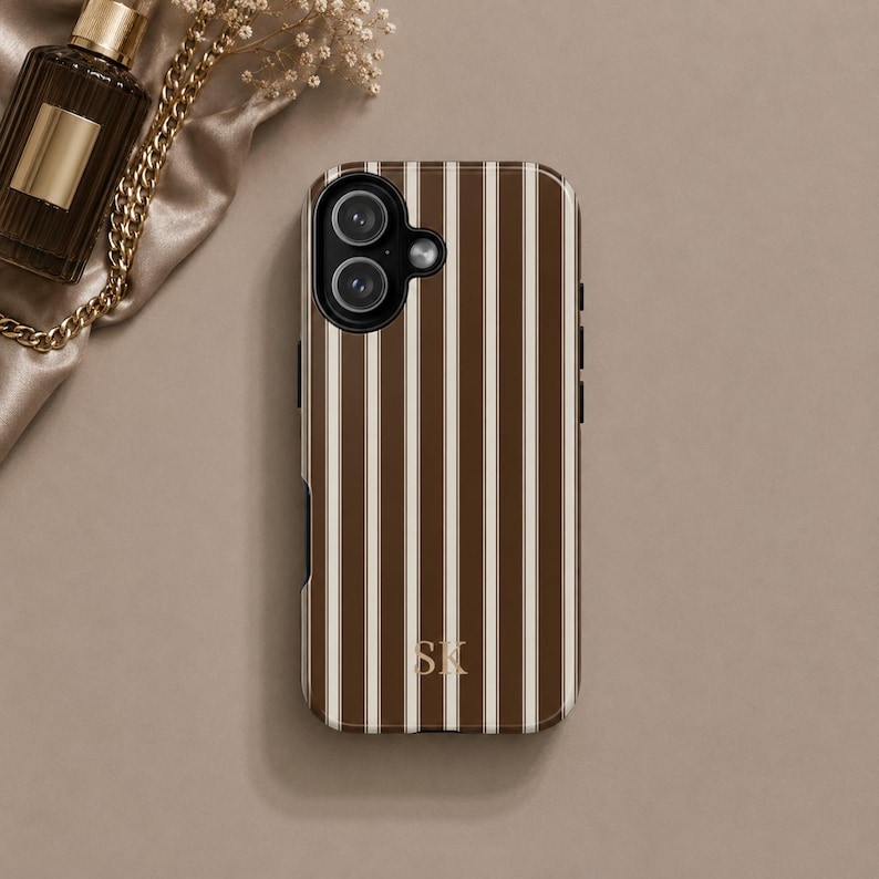K&ouml;nnte beinhalten: Eine Smartphone-H&uuml;lle mit vertikalem Streifenmuster in Dunkelbraun und Creme. Die H&uuml;lle hat oben links einen Ausschnitt f&uuml;r eine Doppelkamera und die goldfarbenen Initialen SK in einer Serifenschrift unten in der Mitte der R&uuml;ckseite.