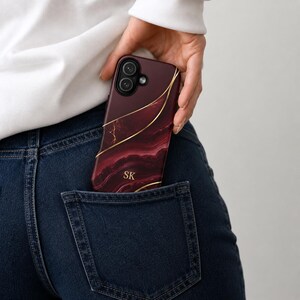 May include: A smartphone case featuring a deep burgundy and dark red marble pattern with thin, flowing gold lines. The case includes the gold-colored initials SK printed on the lower section and has a cutout for a dual-lens camera system.