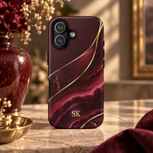 May include: A burgundy phone case featuring a swirling marble pattern with thin gold lines running diagonally across the surface. The letters SK are printed in gold near the bottom center. The case has a circular cutout for a dual camera system.