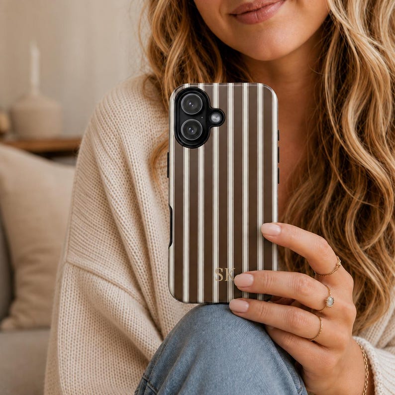 K&ouml;nnte beinhalten: Eine Smartphone-H&uuml;lle mit vertikalem Streifenmuster in abwechselnden T&ouml;nen von Dunkelbraun und Creme. Die H&uuml;lle verf&uuml;gt &uuml;ber einen kreisf&ouml;rmigen Kameraausschnitt und die goldfarbenen Initialen SK im unteren Bereich der R&uuml;ckseite.