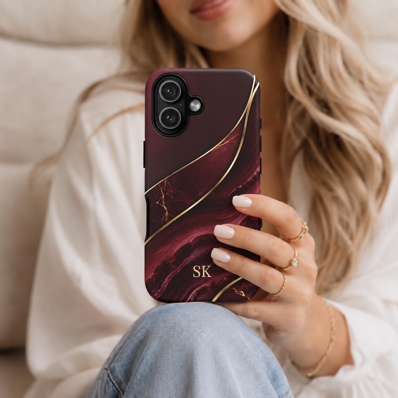 May include: A smartphone case featuring a deep burgundy and black marble pattern with gold-colored wavy lines running diagonally across the surface. The letters SK are printed in gold near the bottom center of the case, which has a dual camera cutout.