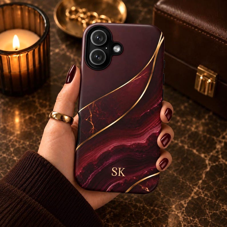 May include: A dark burgundy and black marble-patterned phone case featuring thin, sweeping gold lines and the gold-colored initials SK printed near the bottom. The case has a cutout for a dual-lens camera system and a flash on the upper left side.