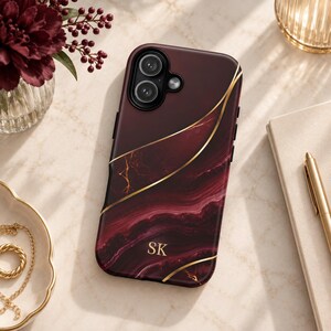May include: A burgundy phone case with a dark red marble pattern and gold-colored vein accents. The case features a dual-lens camera cutout and the gold-colored initials SK printed in the lower center. The design includes curved gold lines.