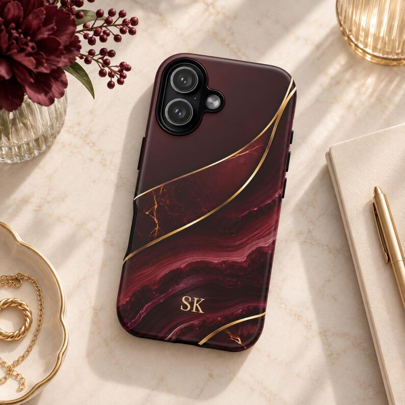 May include: A burgundy phone case with a dark red marble pattern and gold-colored vein accents. The case features a dual-lens camera cutout and the gold-colored initials SK printed in the lower center. The design includes curved gold lines.