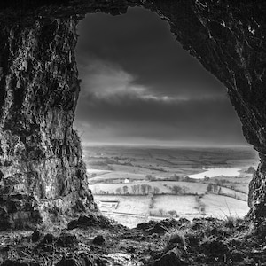 May include: Black and white landscape photograph framed by a dark, textured rock formation. The view reveals a valley with fields, trees, and a body of water under a cloudy sky. The image captures a natural scene.