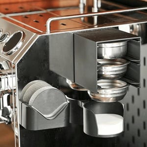May include: A stainless steel espresso machine accessory organizer featuring a black matte finish. It includes a vertical rack for three metal portafilter baskets, a side holder for two circular mesh puck screens, and a lower cup for a white filter disc.