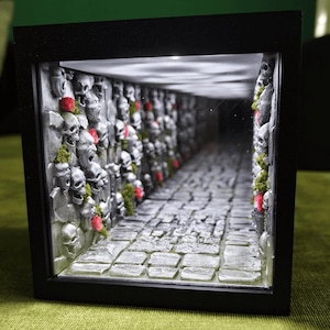 May include: A black-framed diorama featuring a stone-textured corridor lined with silver-painted skulls and small green moss accents. The walls are adorned with tiny red roses, and the floor consists of rectangular stone tiles, illuminated by overhead lighting.