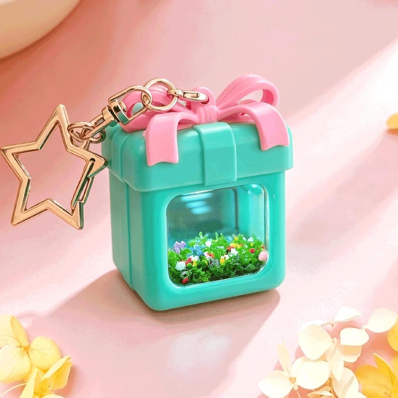 May include: A teal square gift box keychain with a pink bow on top and a clear window on the front. Inside the window is a miniature garden scene with green foliage and tiny colorful flowers. A gold star-shaped metal clasp is attached to the top.