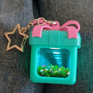 May include: A turquoise gift box keychain with a pink bow on top and a star-shaped metal clasp. The center of the box features a clear window revealing a miniature display of green moss-like material with small colorful accents inside the container.