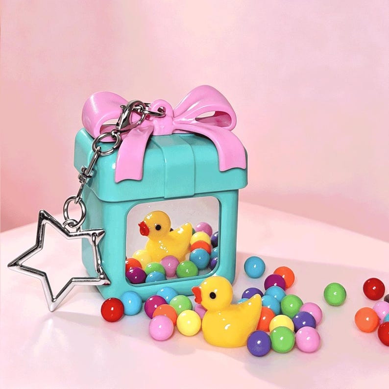 May include: A turquoise gift box-shaped keychain with a pink bow on top and a clear window displaying a yellow rubber duck inside. A silver star-shaped charm is attached to the metal ring. Small, colorful round beads are scattered around the base.